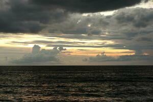 A cloudy sky over the ocean with a dark sky and the sun setting behind it photo