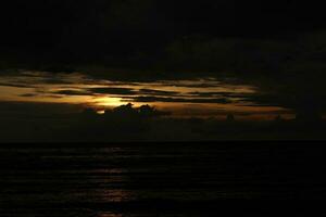 A cloudy sky over the ocean with a dark sky and the sun setting behind it photo