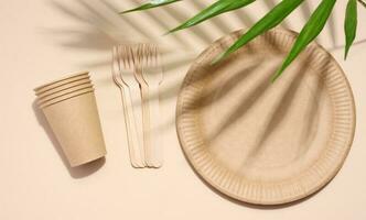 Brown paper plate, stack of glasses and wooden fork, disposable tableware on beige background, top view photo