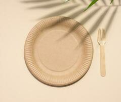 Brown paper plate and wooden fork, disposable tableware on a beige background, top view photo