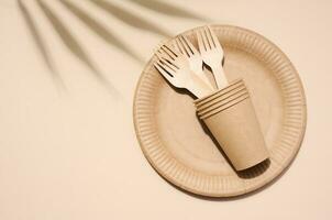 Brown paper plate, stack of glasses and wooden fork, disposable tableware on beige background, top view photo