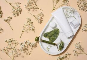 White plastic tube, jade gouache massager and massage jade roller on a beige background photo