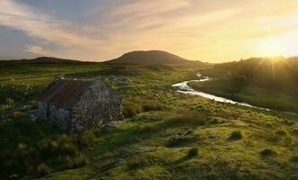 Beautiful sunset scenery of old rusty tin roof cottage on green hill by the river at Connemara National park, county Galway, Ireland photo