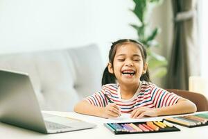 pequeño asiático niña dibujo con color lapices mientras utilizando ordenador portátil a hogar foto