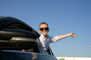A cute boy in sunglasses looked out of the car window waving his hand with a laugh photo