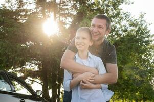 Dad hugs a cute boy standing near the car in the woods, the contour light, the bright sun. photo