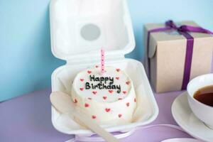 bento cumpleaños pastel en embalaje ecológico con un vela siguiente a un taza de té y un regalo en un púrpura y azul antecedentes foto