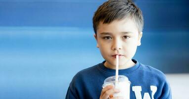 Kid Drinking Healthy Juice,Cute young boy using straw drinking summer fruit smoothie from plastic glass on blue background with copy space,Portrait School Kid loooking at Camera photo