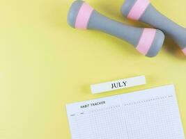 flat lay of habit tracker book, wooden calendar July, gray pink dumbbells on yellow  background with copy space. Self development and active lifestyle concept. photo