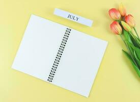 flat lay of opened blank pages notebook or diary, wooden calendar July with tulips bouquet on yellow background  with copy space. photo