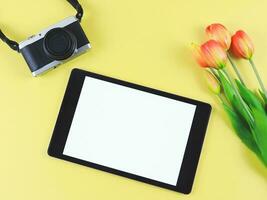 flat lay of digital tablet with blank white screen, tuip flowers  and digital camera isolated on yellow background. photo