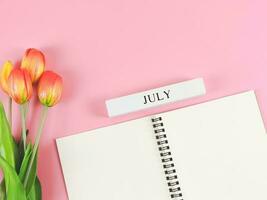 flat lay of opened diary or notebook with wooden calendar july  on pink background with orange-yellow tulips. photo