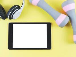 flat lay of digital tablet with blank white screen,  two pink grey dumbbells and headphones  isolated on yellow background. Online training concept. photo