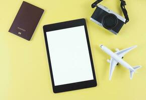 flat lay of digital tablet with blank white screen, airplane model, passport and digital camera isolated on yellow background. travel planning concept. photo