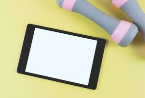 flat lay of digital tablet with blank white screen and two pink grey dumbbells  isolated on yellow background. photo