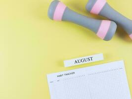 flat lay of habit tracker book, wooden calendar August, gray pink dumbbells,  on yellow  background with copy space. Self development and active lifestyle concept. photo