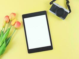 plano laico de digital tableta con blanco blanco pantalla, tulipán flores y digital cámara aislado en amarillo antecedentes. foto