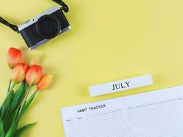 flat lay of habit tracker book, wooden calendar July, digital camera and red yellow tulips flowers on yellow background with copy space. photo