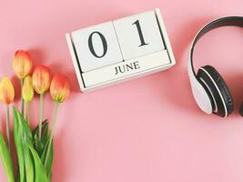 flat lay of wooden calendar with date June 01, headphones and  orange  yellow tulips on pink background, copy space. photo