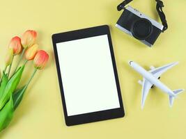 flat lay of digital tablet with blank white screen, tulip flowers, airplane model and  digital camera isolated on yellow background. photo