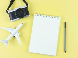 plano laico de blanco página abrió cuaderno con bolígrafo, avión modelo y digital cámara en amarillo antecedentes con Copiar espacio. foto