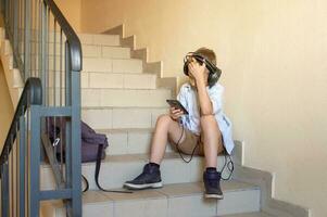 un frustrado chico es sentado en el escalera con un teléfono en su manos y auriculares foto