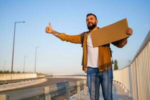 Man is hitchhiking on roadside trying to stop car. He is holding blank cardboard for your text. photo