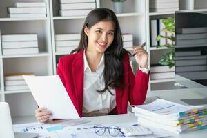 Asian businesswoman in red suit calculating company sales with calculator, laptop, and tablet on table Interior of businesswoman office at the modern workstation. photo