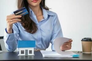 Asian woman with long hair in the blue shirt is using a credit card for buying a new house and looking at home-buying documents The idea of purchasing a home with a credit card. photo