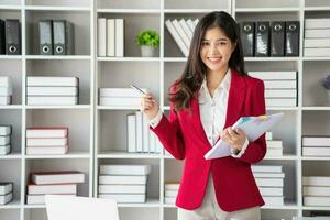 Asian businesswoman in red suit calculating company sales with calculator, laptop, and tablet on table Interior of businesswoman office at the modern workstation. photo