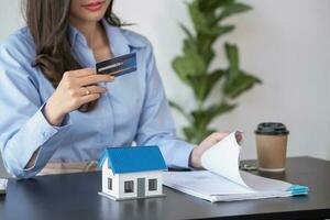 asiático mujer con largo pelo en el azul camisa es utilizando un crédito tarjeta para comprando un nuevo casa y mirando a comprar casa documentos el idea de adquisitivo un hogar con un crédito tarjeta. foto