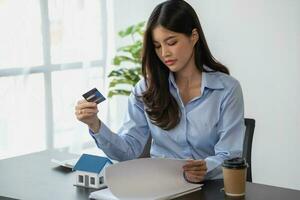 asiático mujer con largo pelo en el azul camisa es utilizando un crédito tarjeta para comprando un nuevo casa y mirando a comprar casa documentos el idea de adquisitivo un hogar con un crédito tarjeta. foto