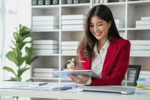 Asian businesswoman in red suit calculating company sales with calculator, laptop, and tablet on table Interior of businesswoman office at the modern workstation. photo