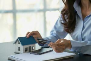 asiático mujer con largo pelo en el azul camisa es utilizando un crédito tarjeta para comprando un nuevo casa y mirando a comprar casa documentos el idea de adquisitivo un hogar con un crédito tarjeta. foto