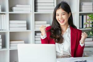 Happy Asian businesswoman in red suit showing happy gesture holding smartphone at desk Portrait of beautiful smiling businesswoman working at a modern workstation. photo