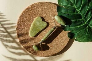 Jade Gua sha scraper and face roller massager on a cork round stand with a monstera leaf. photo