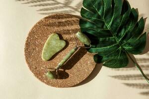 Jade Gua sha scraper and face roller massager on a cork round stand with a monstera leaf. photo