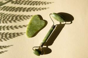 Jade Gua sha scraper and face roller massager on a cork round stand with a monstera leaf. photo