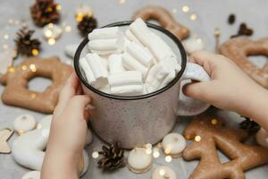 para niños manos sostener un taza de cacao con Malvaviscos. Navidad pan de jengibre en el mesa, bokeh luces en el primer plano. el concepto de postres y bebidas durante el Navidad vacaciones. foto