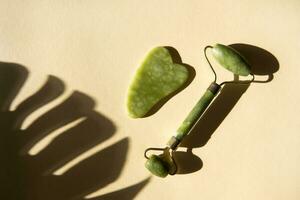 Jade Gua sha scraper and face roller massager on a cork round stand with a monstera leaf. photo