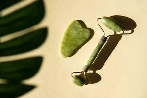 Jade Gua sha scraper and face roller massager on a cork round stand with a monstera leaf. photo