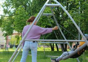 Sankt Petersburgo Rusia - 06 10 2023 asiático madre ayuda a empujar columpios para dos pequeño niña tener divertido en patio de recreo de público parque. foto