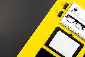 Modern office desk flat lay with spiral paper notepad, white envelopes mockup and reading glasses on black yellow background. Top view. Copy space. photo