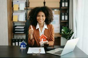 Young real estate agent worker working with laptop and tablet at table in office and small house beside it. photo