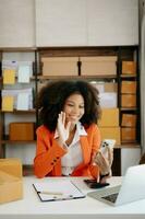 puesta en marcha pequeño negocio pequeño, empresario propietario africano mujer utilizando teléfono inteligente o tableta tomando recibir y comprobación en línea compra compras orden a preparando paquete producto caja. foto