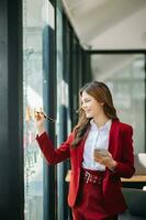 secretario trabajando en moderno oficina en rascacielos, escritura y pega adhesivo notas con Tareas en ventana oficina foto