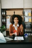 Young real estate agent worker working with laptop and tablet at table in office and small house beside it. photo