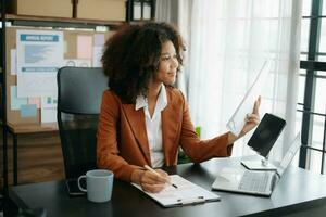 confidente hermosa africano mujer de negocios mecanografía ordenador portátil computadora y digital tableta mientras participación café a oficina foto