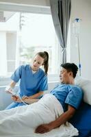 Friendly Female Head Nurse Making Rounds does Checkup on Patient Resting in Bed. She Checks tablet while Man Fully Recovering after Successful Surgery in hospital photo