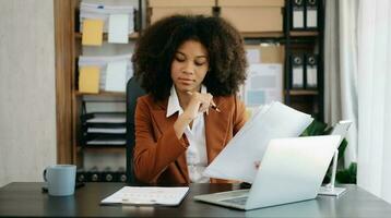 Confident beautiful African businesswoman typing laptop computer and digital tablet while holding coffee at office photo